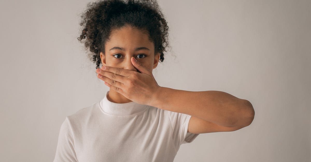 Who held the girl in the secret room? - Concerned African American teenage girl in white outfit looking at camera while covering mouth with hand on gray background in studio