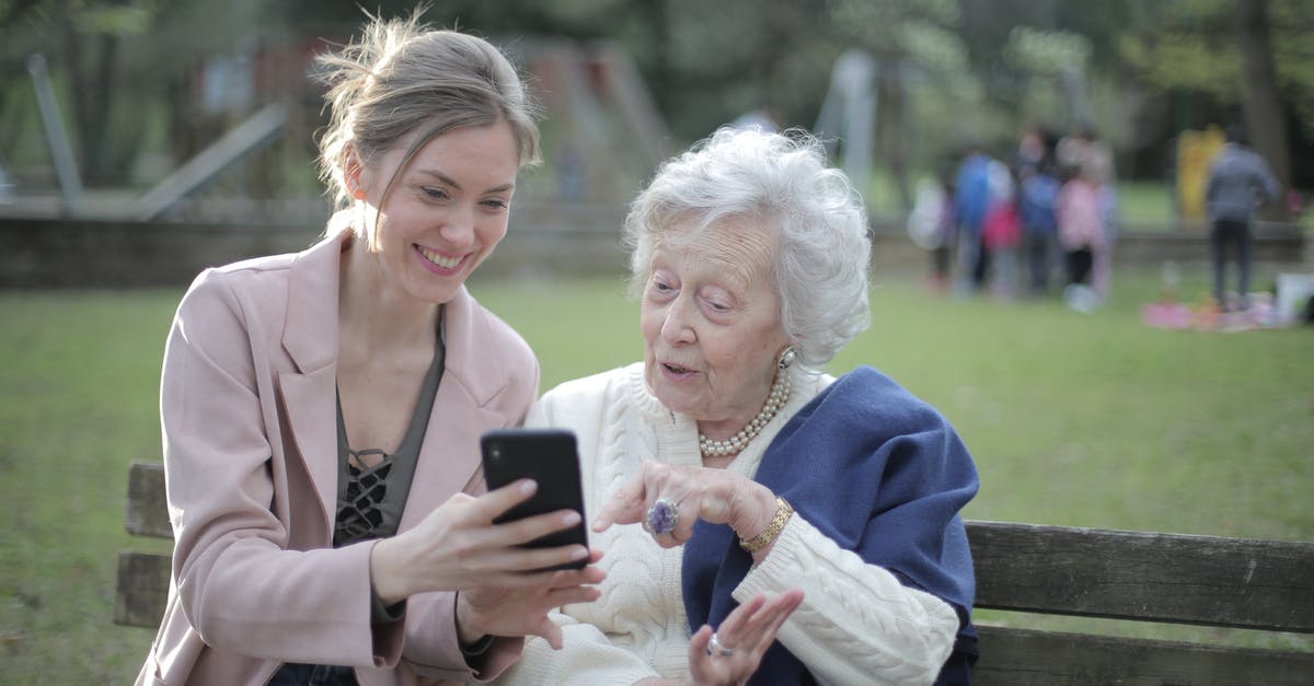Who is elder in Charlie and Alan? - Delighted female relatives sitting together on wooden bench in park and browsing mobile phone while learning using