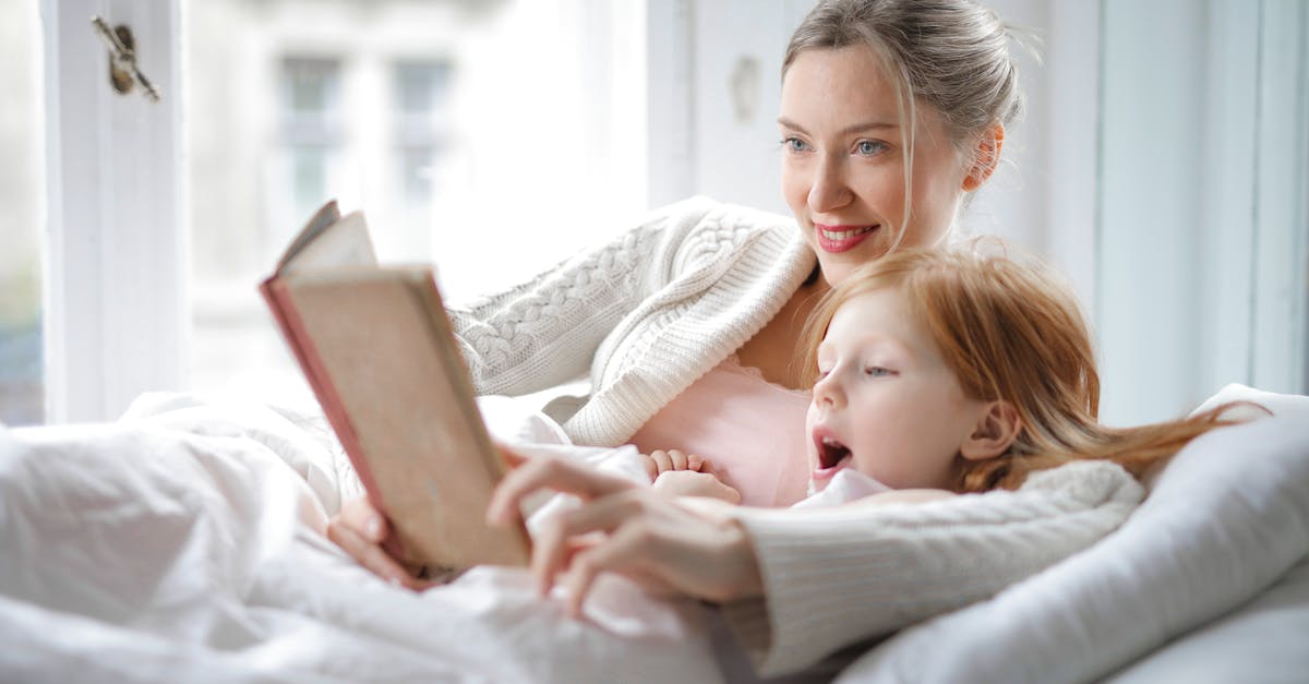 Who is Katniss' love interest? - Cheerful young woman hugging cute little girl and reading book together while lying in soft bed in light bedroom at home in daytime