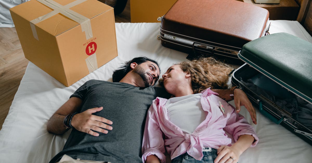 Who threw the suitcase to the other? - From above of young ethnic bearded man and woman with curly hair looking at each other and lying on bed among suitcases and cardboard boxes with stuff while moving in new apartment together