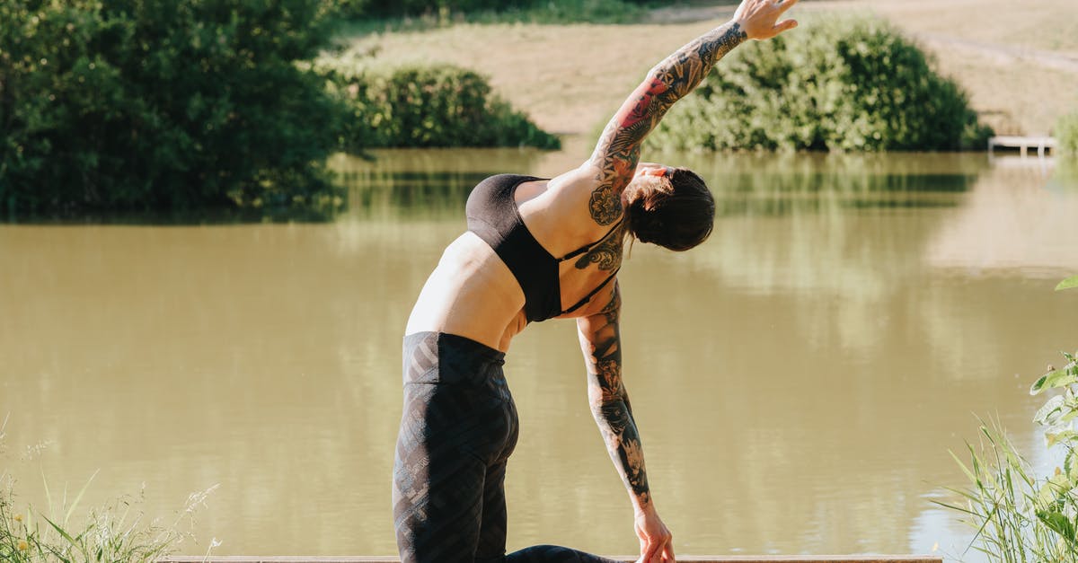 Who was the body in the lake? - Side view of unrecognizable flexible tattooed lady in sports clothes standing on knees with raised arm on dock against river