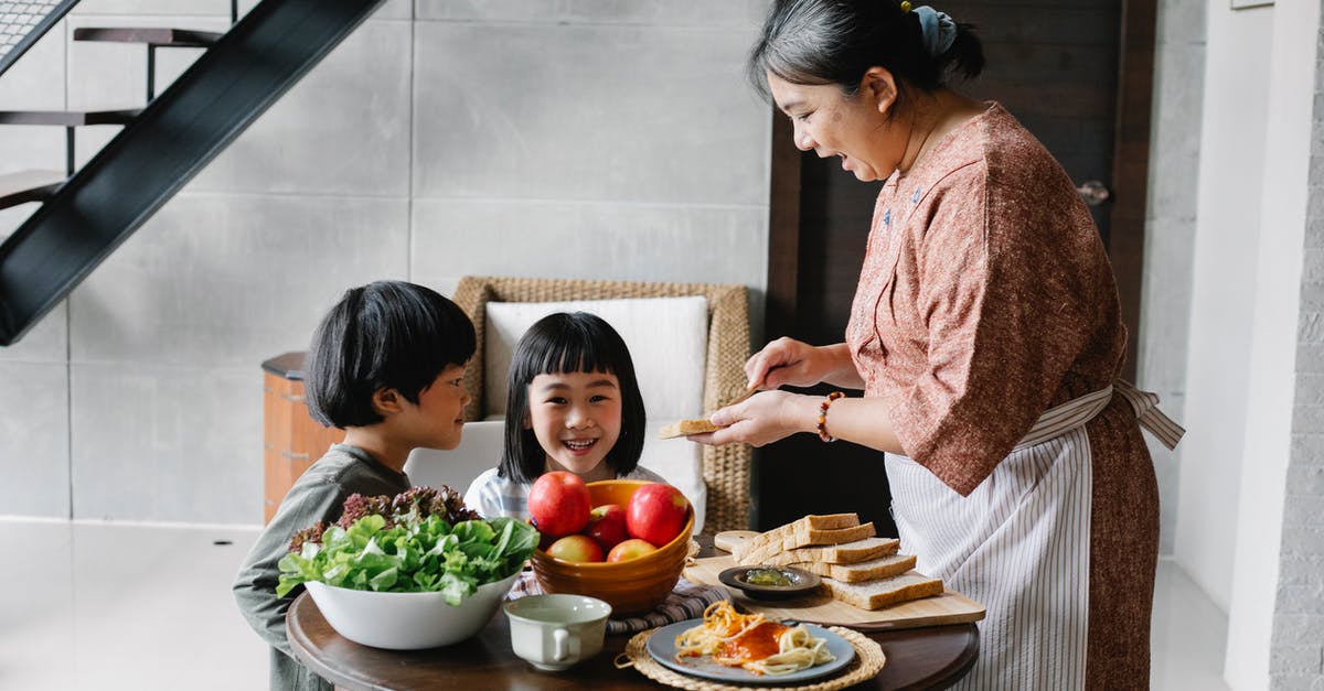 Why did Gordon make Evey the same breakfast and also saying bonjour to her? - Side view of positive senior Asian female in apron spreading butter on bread while preparing breakfast for funny little grandchildren sitting at round table in cozy kitchen