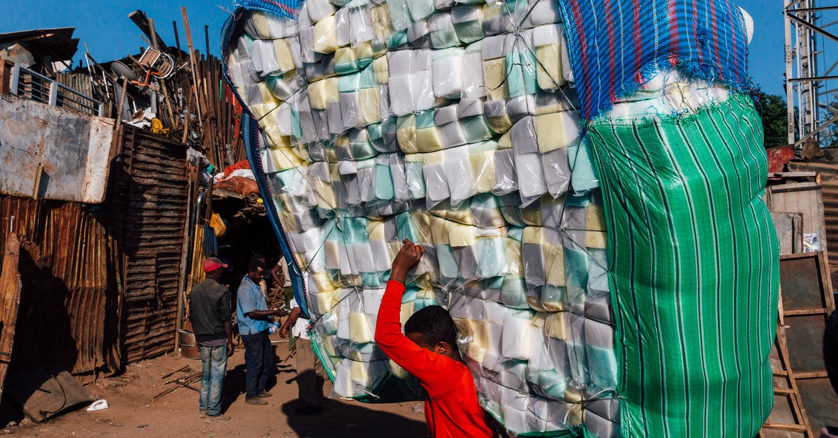Why did the Big Guy say "Small world" when she first arrived? - Side view of anonymous ethnic male in casual wear carrying heavy stack of merchandise with striped bags on dry terrain under blue sky in sunlight