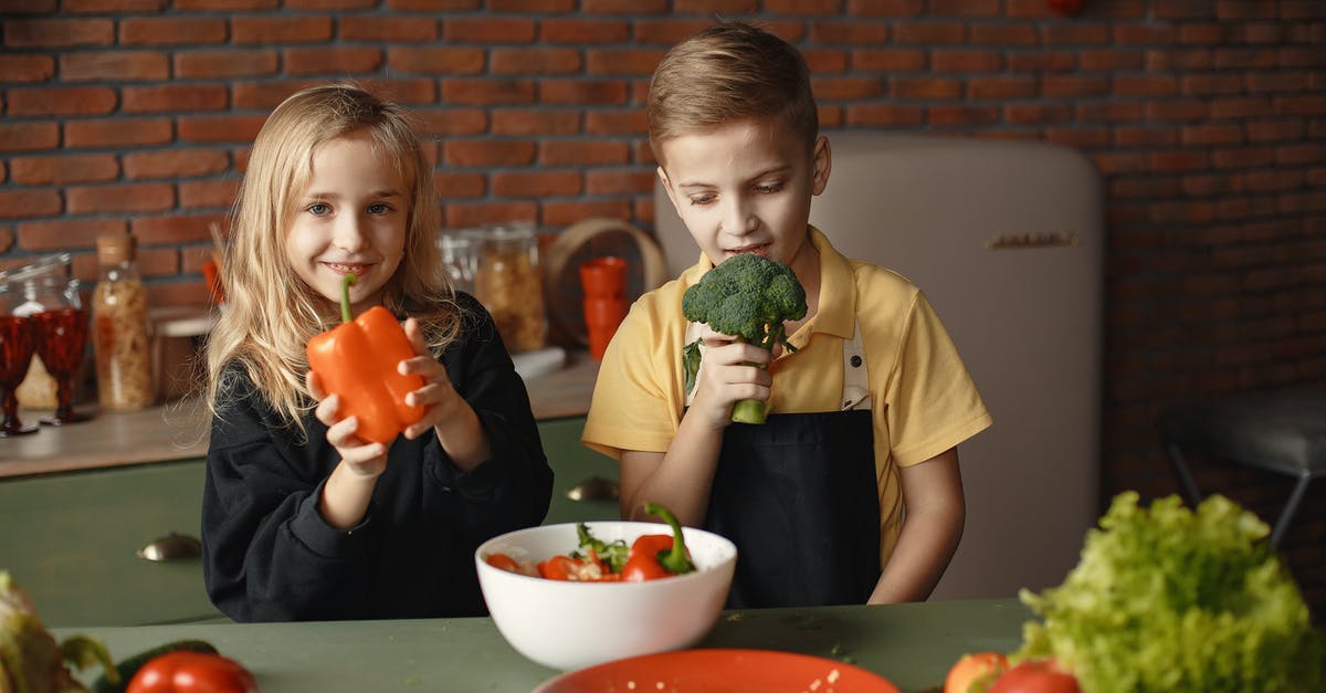 Why didn't he take the kids? - Cute girl and boy with fresh pepper and broccoli in hands standing near desk with assorted vegetables and salad bowl during vegetarian meal preparation