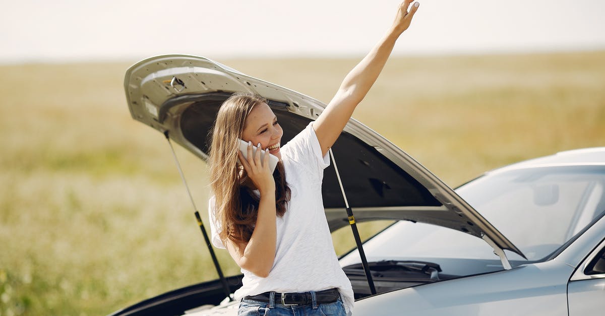 Why didn't they call for this character's help sooner? - Cheerful young female driver in white t shirt and jeans speaking on phone and looking away with smile while leaning on broken car with open hood and showing greeting gesture with raised hand