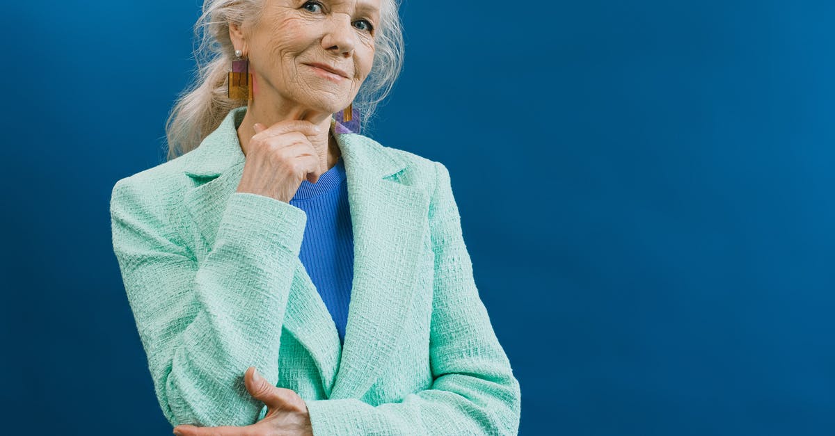 Why do actors in old movies look old? - Elderly smiling gray haired female wearing mint blazer and earnings standing against blue background touching chin and looking at camera