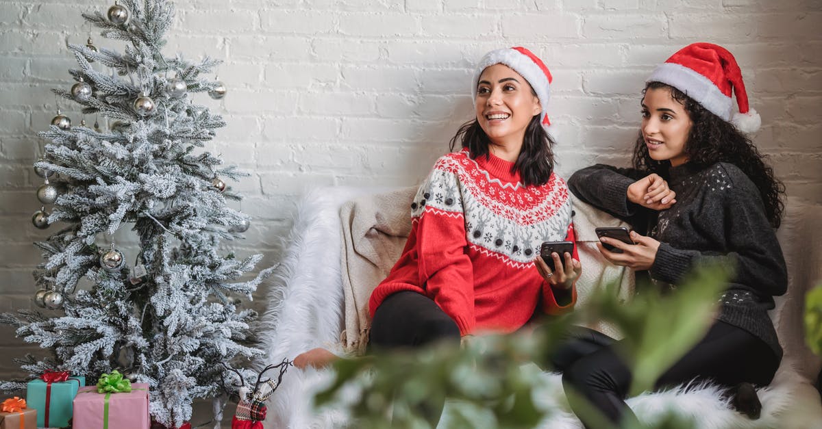 Why do Adam and Eve live so far apart? - Happy young Hispanic women in Santa hat with mobile phones sitting on small sofa near decorated Christmas tree and looking away