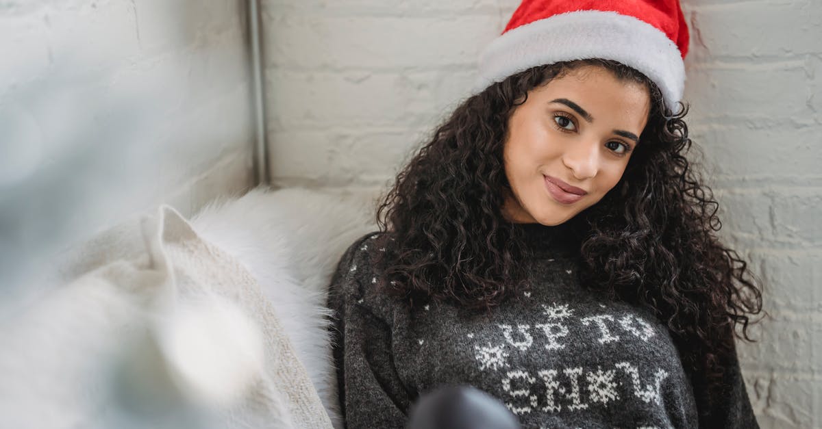 Why do Adam and Eve live so far apart? - Young female in warm sweater and hat sitting on sofa and looking at camera while resting in cozy room on Christmas