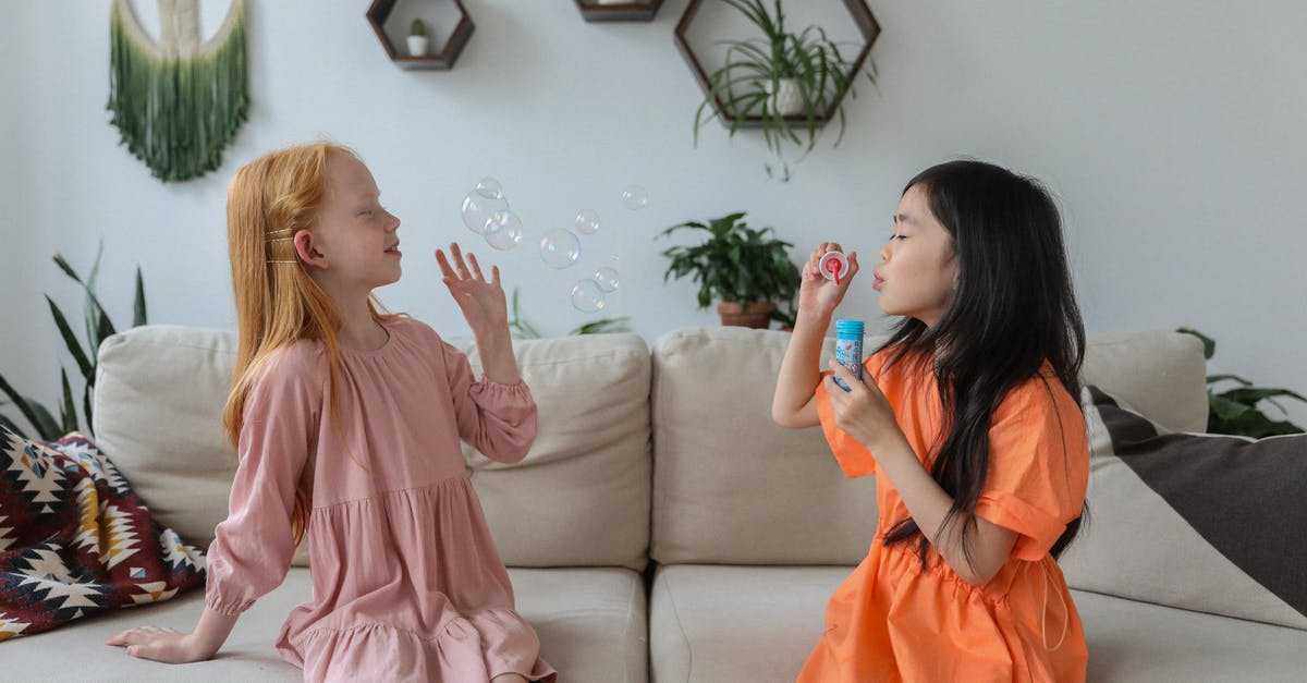 Why do soap operas have the soap opera effect? - Side view of cheerful little diverse girlfriends in trendy dresses sitting on comfortable sofa in cozy living room and blowing soap bubbles