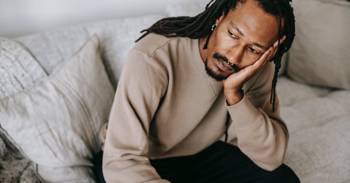 Why does Guy consider changing sex? - Pensive African American male with dreadlocks in casual clothes sitting on comfortable couch and pondering touching face