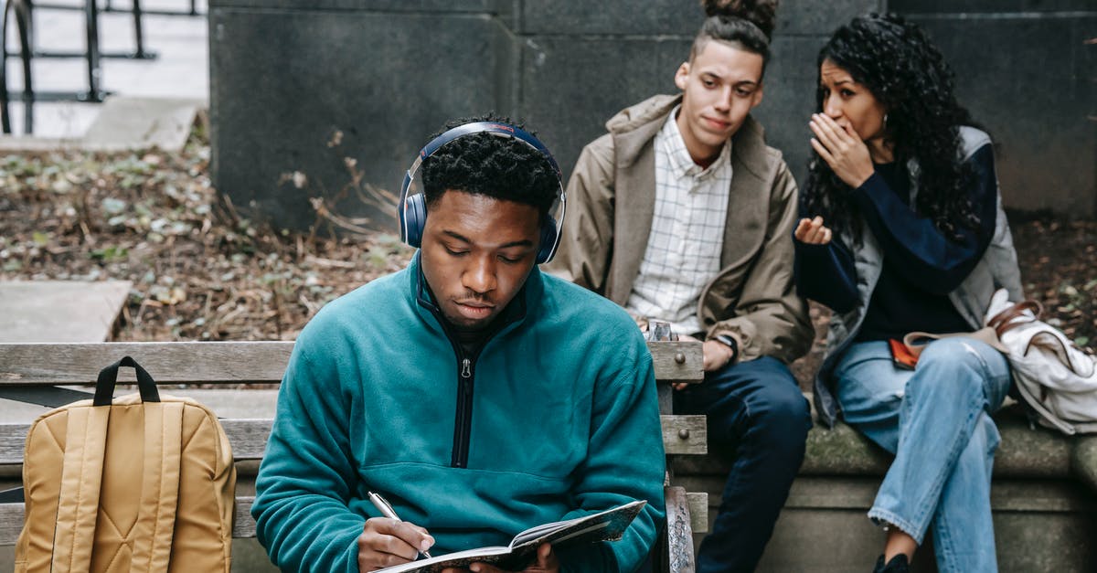 Why does Indy pause while writing "neolithic" in the campus scene? - Young multiethnic friends in casual outfit with backpack gossiping behind back of African American guy sitting on bench in headphones and taking notes in notebook in city street in daylight