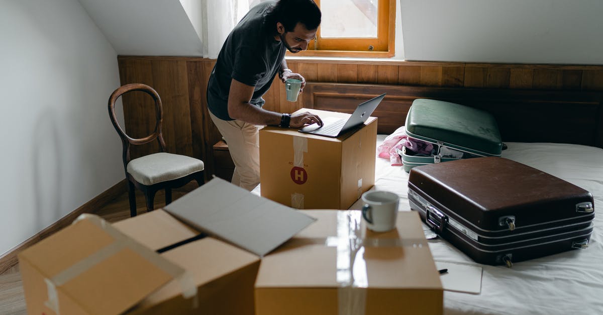 Why does the chair move by itself? - Side view of ethnic male in casual clothes and wristwatch surfing internet on netbook and standing with cup of hot drink surrounded by cardboard boxes and suitcases in bedroom