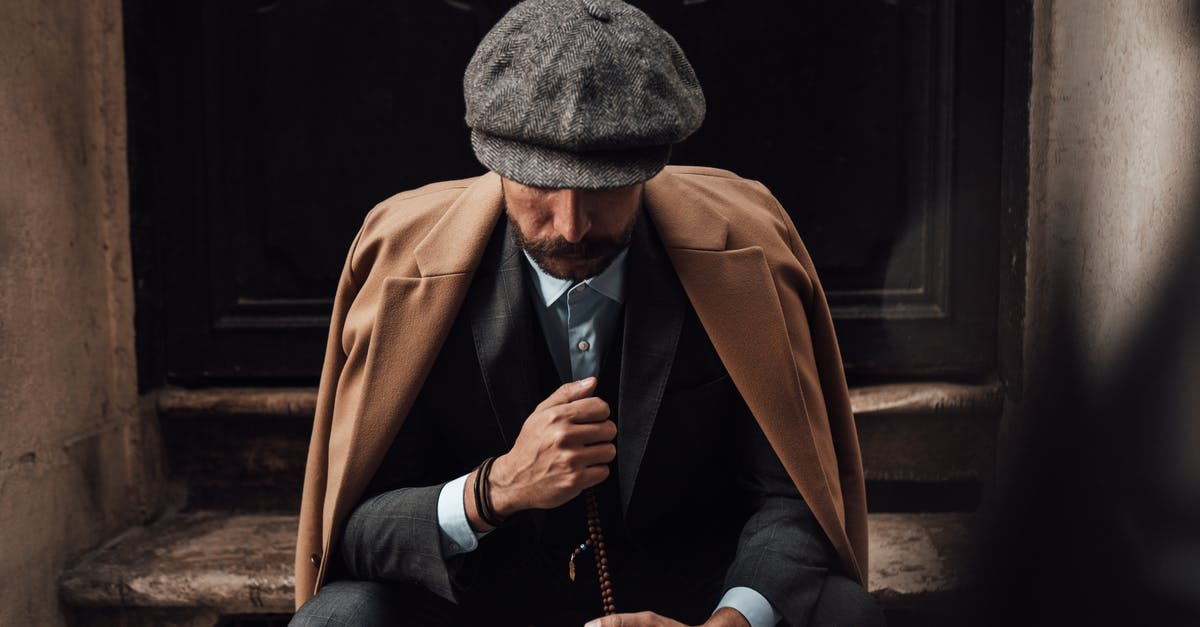 Why does the Dude look like he does? - Pensive stylish male in posh suit coat and hat sitting with beads on shabby doorway and looking down in thoughts