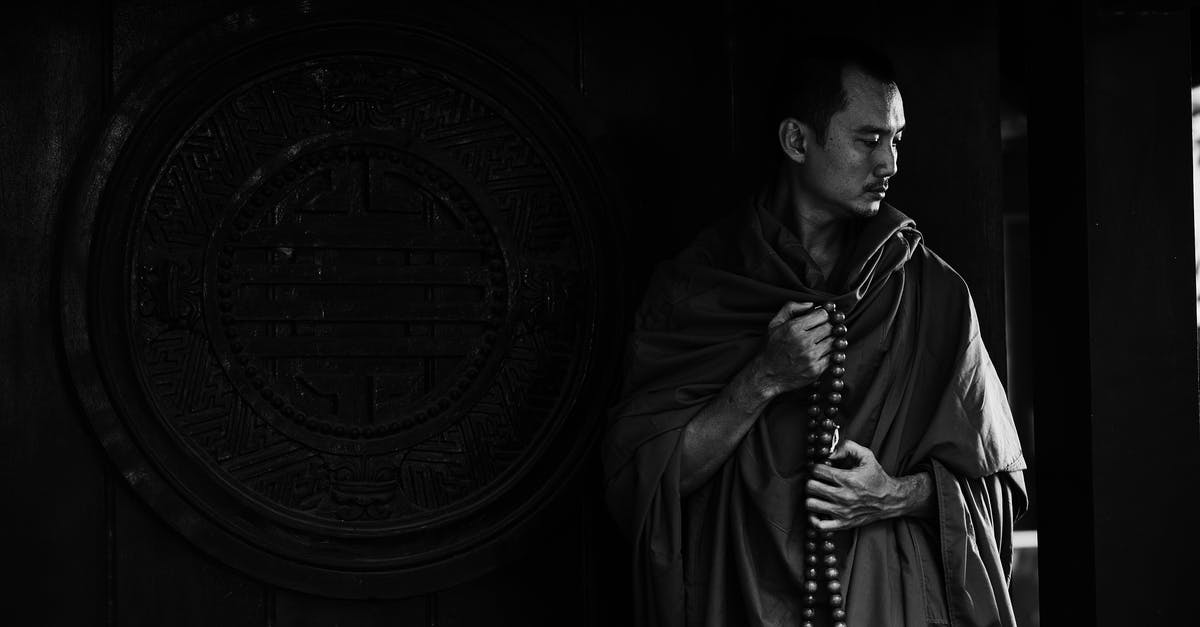 Why does Xerxes think he's a god? - Black and white calm pensive Buddhist monk with beads wearing traditional clothes standing near ornamental temple wall with round caved drawing and looking down in thoughts