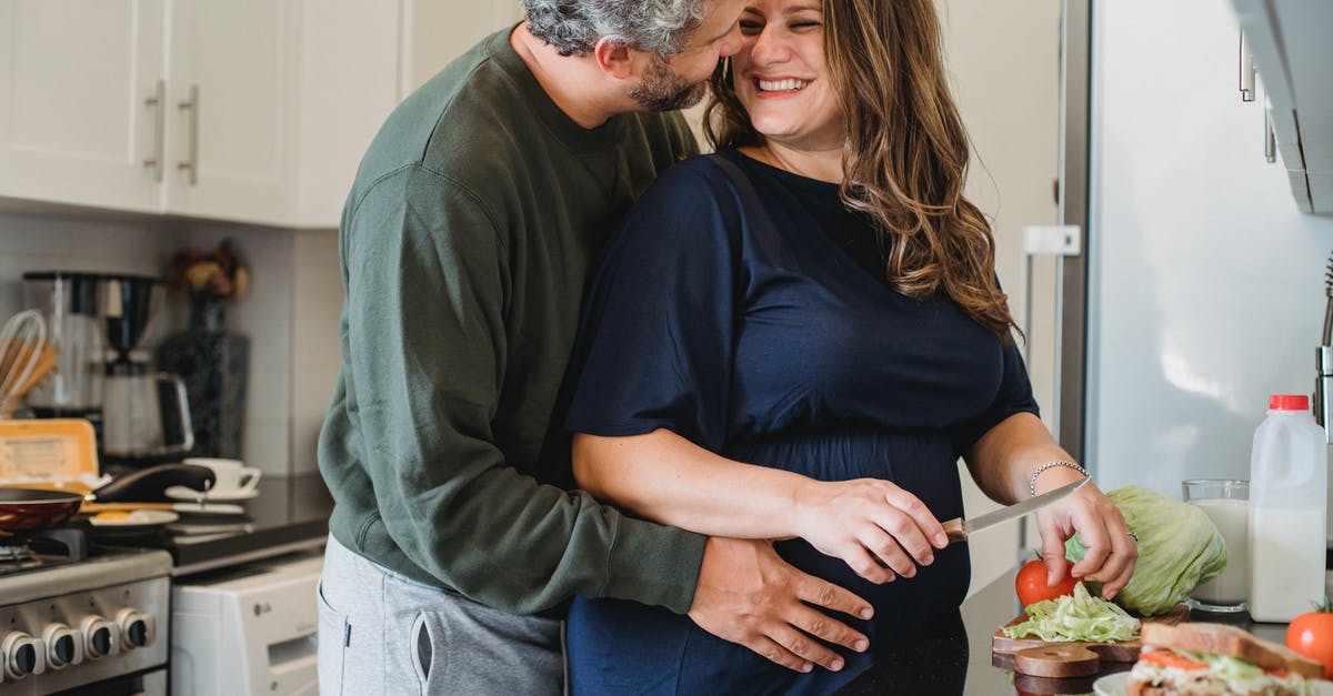 Why doesn't Wayward Pines wait a more couple of thousands of years? - Cheerful pregnant wife in blue dress cooking sandwiches for breakfast while smiling and looking at husband embracing belly in modern kitchen