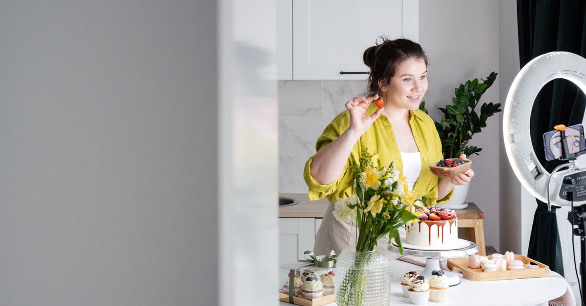 Why is the phone ringing? - Delighted young female influencer in casual clothes smiling and demonstrating fresh berries while decorating appetizing cake during recording vlog on smartphone in kitchen