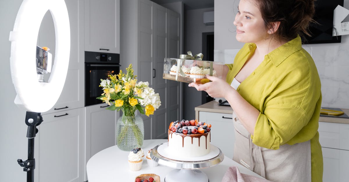 Why is the phone ringing? - Side view of positive young female food blogger in casual clothes and apron introducing yummy cakes and smiling while standing in front of ring lamp and recording vlog on smartphone