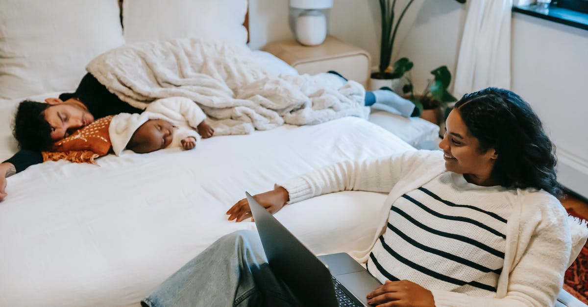 Why the movie title “The Big Sleep?” - High angle of joyful young ethnic mother smiling while watching movie on netbook sitting near adorable baby sleeping on bed with father during weekend at home